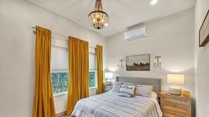 Une chambre à coucher avec un lit, des rideaux, un lustre, une table de chevet et un climatiseur fixé au mur.