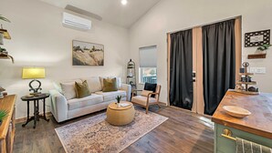 Un salon moderne avec un canapé blanc, une table basse, une table d’appoint, un tapis, un fauteuil, une fenêtre avec des rideaux, un tableau, un climatiseur mural et une petite étagère avec des plantes.