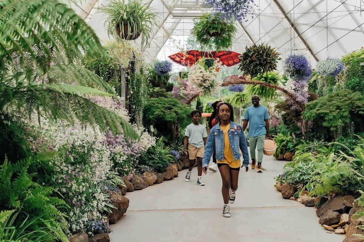 Een kas met verschillende planten en bloemen, een pad en mensen die wandelen.