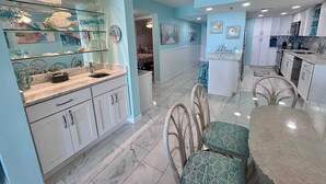 Una cocina amplia con armarios blancos, un comedor con mesa de vidrio y una sala de estar con cama y detalles decorativos en la pared.