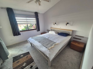 Une chambre à coucher comprenant un lit, des tables de chevet, une fenêtre avec des rideaux, un ventilateur de plafond et une vue sur l’extérieur.