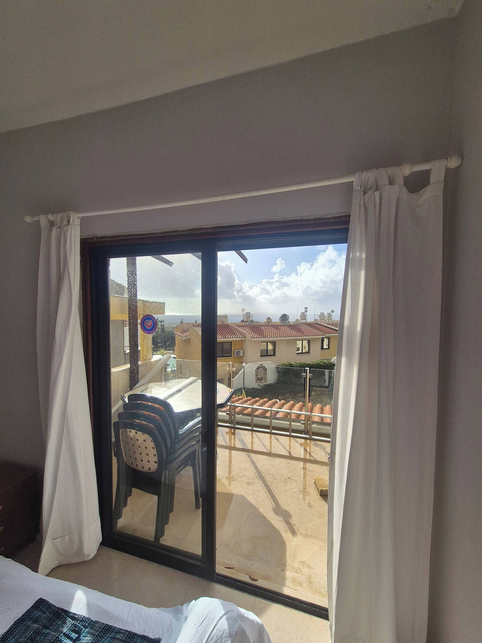 Ein Zimmer mit einer großen Schiebeglastür, die auf einen Balkon führt. Der Balkon ist mit einem Metallgeländer versehen und bietet Blick auf die Straße und die Häuser.