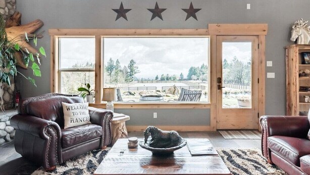A spacious living room with a large window, leather sofas, a coffee table, and a decorative horse sculpture. - Roosters Coop - Wine Country at a Rustic, Lodge Style Retreat with Hot Tub & Pool (Newberg)
