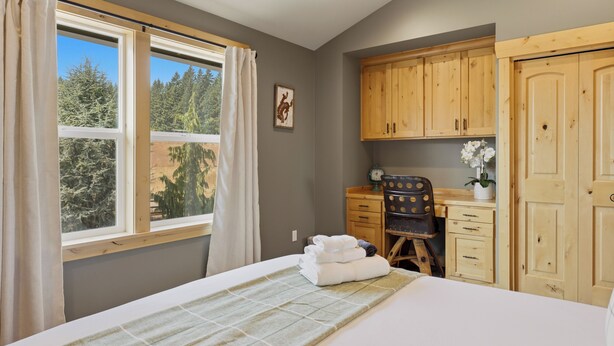 Ein Schlafzimmer mit einem Bett, einem Schreibtisch mit Stuhl, Holzschränken, einem Fenster mit Vorhängen und Blick auf Bäume.