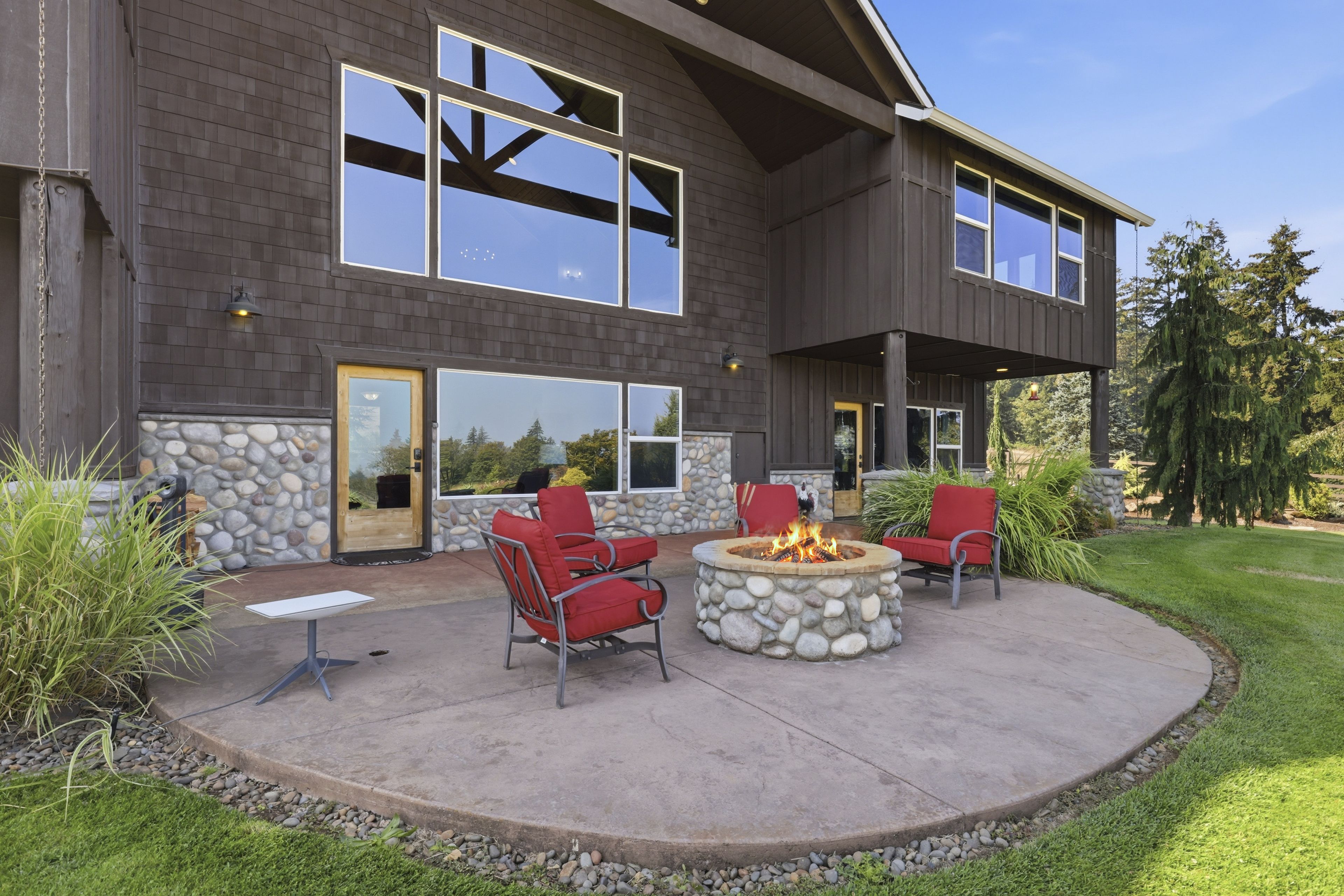 A two-story house with a stone facade, large windows, and a stone fire pit with red chairs.