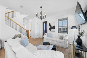A modern living room with a staircase, white sofas, a chandelier, and a large window with blinds. - Family Retreat-Walk to Beach-King Beds-Work Space-Golf Cart (Orange Beach)