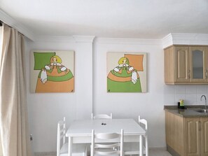 A small dining area with a white table and chairs, two paintings on the wall, and a kitchen area with wooden cabinets.