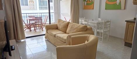 A living room with a sofa, a chair, a TV, and a balcony with a table and chairs.