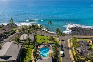 An aerial view of a residential area with a swimming pool surrounded by palm trees, a road, and a view of the ocean. - Nihi Kai Villas 700 Ocean Views AC (Koloa)