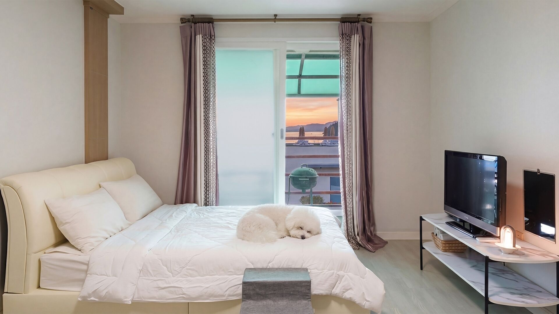 Ein Schlafzimmer mit Bett, Fernseher und Blick auf einen Sonnenuntergang durch ein Fenster.