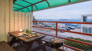 Een balkon met een houten tafel gedekt voor een maaltijd, een barbecue met eten en uitzicht op een kustgebied met gebouwen en auto's.