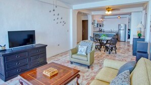 A living room with a TV, sofa, and coffee table. There is a dining area with a table and chairs. The kitchen is visible in the background. - Department Pino Amapas (Puerto Vallarta)