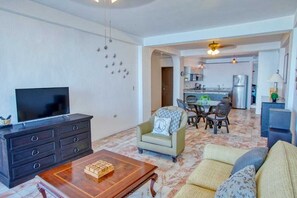 A living room with a TV, sofa, and coffee table. There is a dining area with a table and chairs. The kitchen is visible in the background. - Department Pino Amapas (Puerto Vallarta)
