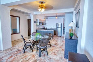 A dining area with a glass table and chairs, a kitchen in the background, and a ceiling fan. - Department Pino Amapas (Puerto Vallarta)