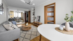 A modern living room with a grey sofa, a round coffee table, and a white dining table. There is a wooden door with glass panels, a ceiling fan, and a plant on the dining table. - Mokotów Urban Cozy Apartment (Warszawa)