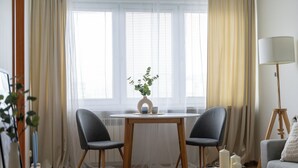 A cozy living room with a round wooden table, two grey chairs, a white lamp, and a window with sheer curtains. - Mokotów Urban Cozy Apartment (Warszawa)