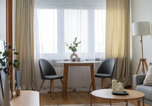 Ein gemütliches Wohnzimmer mit einem runden Holztisch, zwei grauen Stühlen, einer weißen Lampe und einem Fenster mit durchsichtigen Vorhängen.