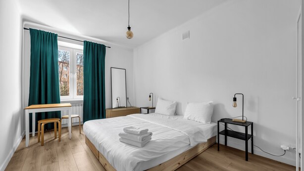 Ein modernes Schlafzimmer mit einem hölzernen Bett, weißer Bettwäsche, einem Schreibtisch mit Stuhl und einem Nachttisch mit Lampe.