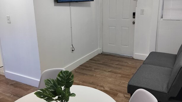 En stue med et hvidt vægmonteret tv, en grå sofa, et rundt hvidt bord med en plante og en fjernbetjening, samt en dør med et vindue.