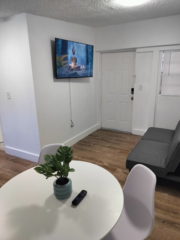 A living room with a white wall-mounted TV, a grey sofa, a round white table with a plant and remote, and a door with a window. - Little Havana Urban Escape  14 (Miami)