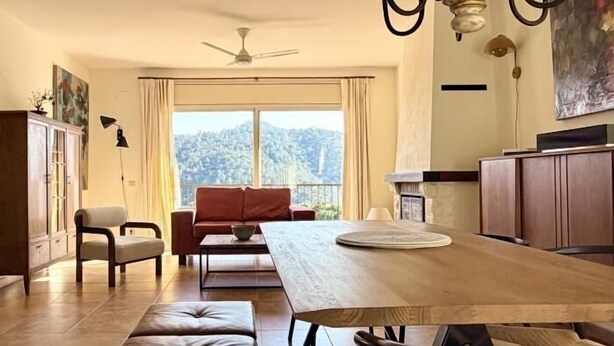 Ein geräumiges Wohnzimmer mit einem großen Holzesstisch, einem roten Sofa und Blick auf eine Berglandschaft durch das Fenster.
