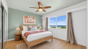 Ein Schlafzimmer mit einem Bett, einem Deckenventilator, einem Fenster mit Blick auf den Strand und einer Wand mit einem Gemälde.