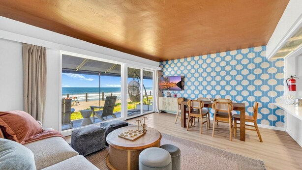Ein Wohnzimmer mit großem Fenster zum Strandblick, einer Essecke mit Holzmöbeln und einer Wand mit Muster.