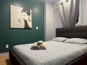Ein Schlafzimmer mit Bett, Kopfteil, Wand mit Gemälde und Vorhang.