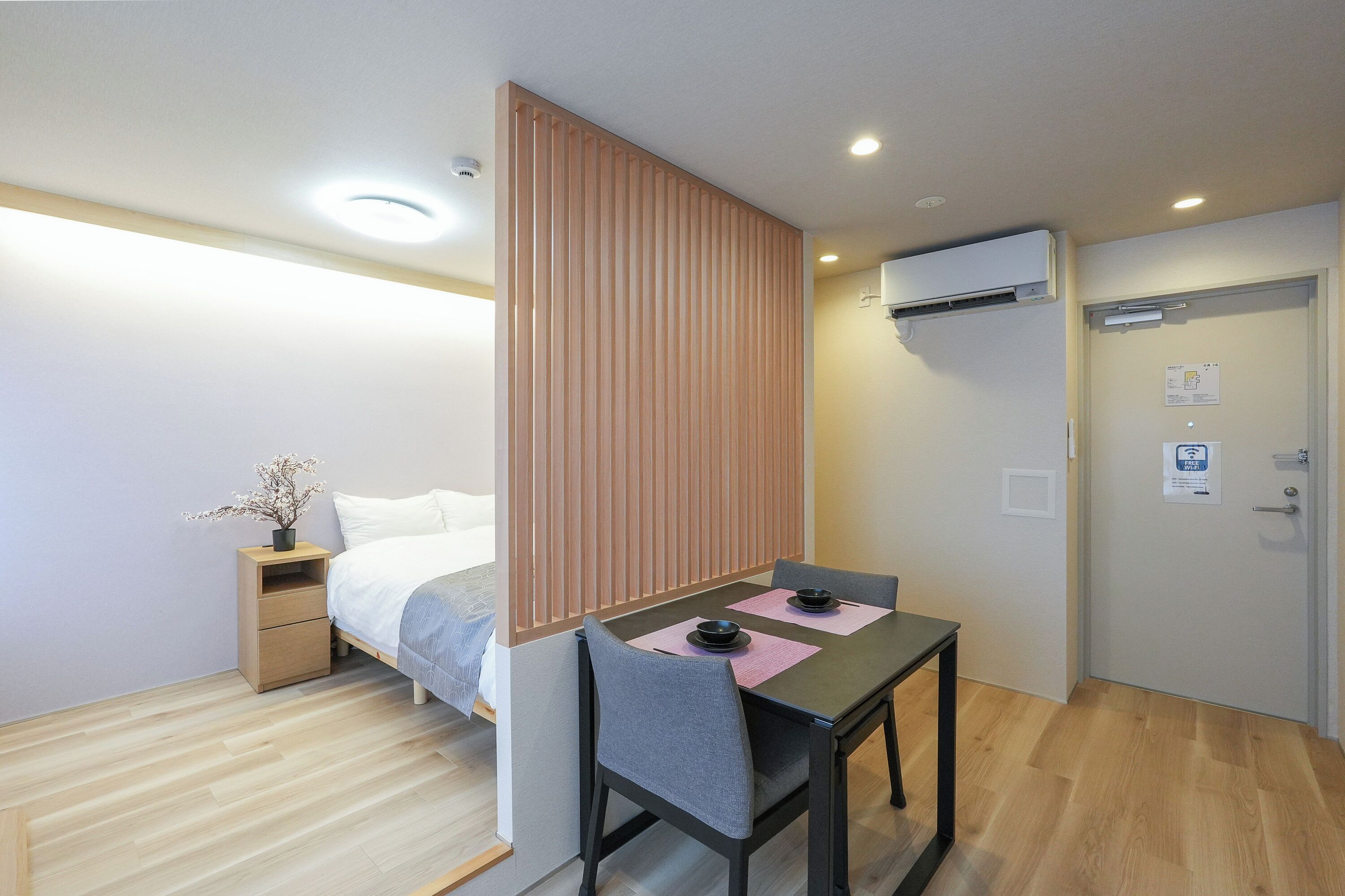 A room with a bed, a table with chairs, a small nightstand, a decorative plant, and a sliding partition.
