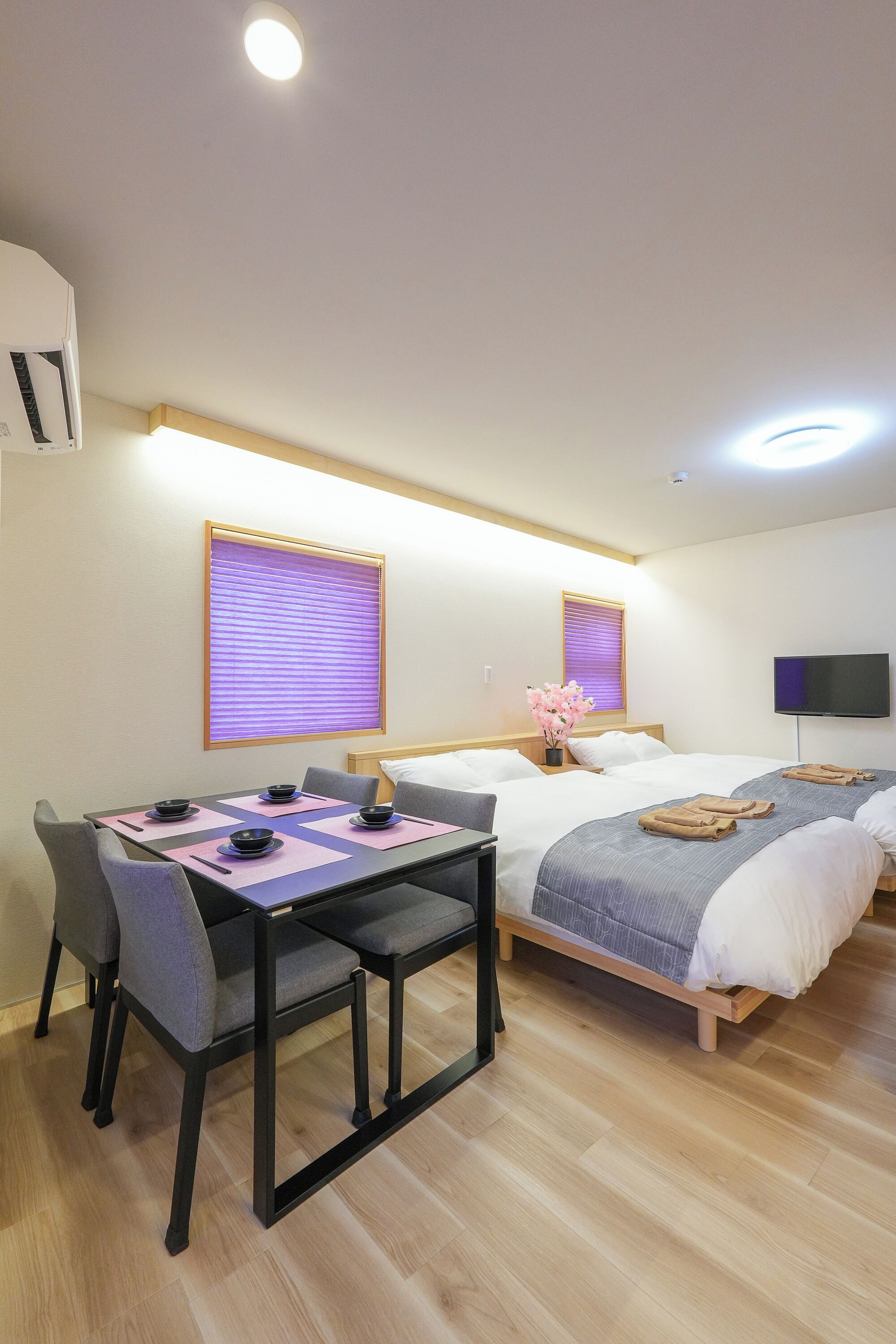 A hotel room with a bed, a TV, a table with chairs, and a window with blinds.