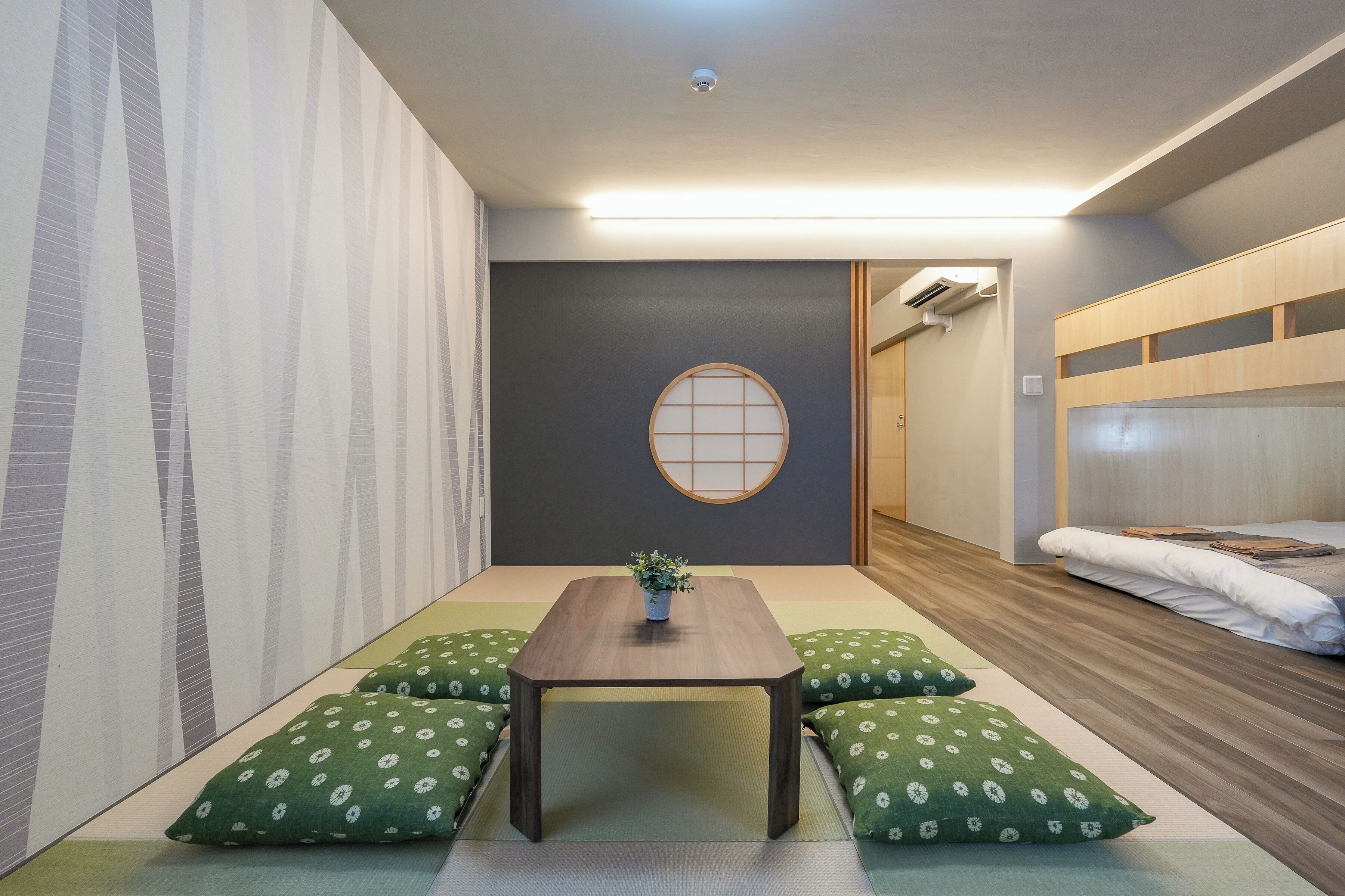 Una habitación con suelo de tatami, una mesa baja de madera, cojines verdes, un panel shoji y una cama con cabecera de madera.