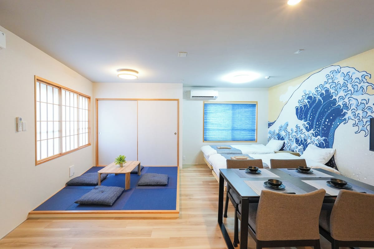 A room with a low platform bed, a dining table with chairs, a small table with a plant, and a large wave mural on the wall.