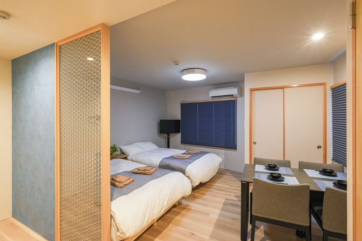 A room with two beds, a dining table with chairs, a window with blinds, and a door.