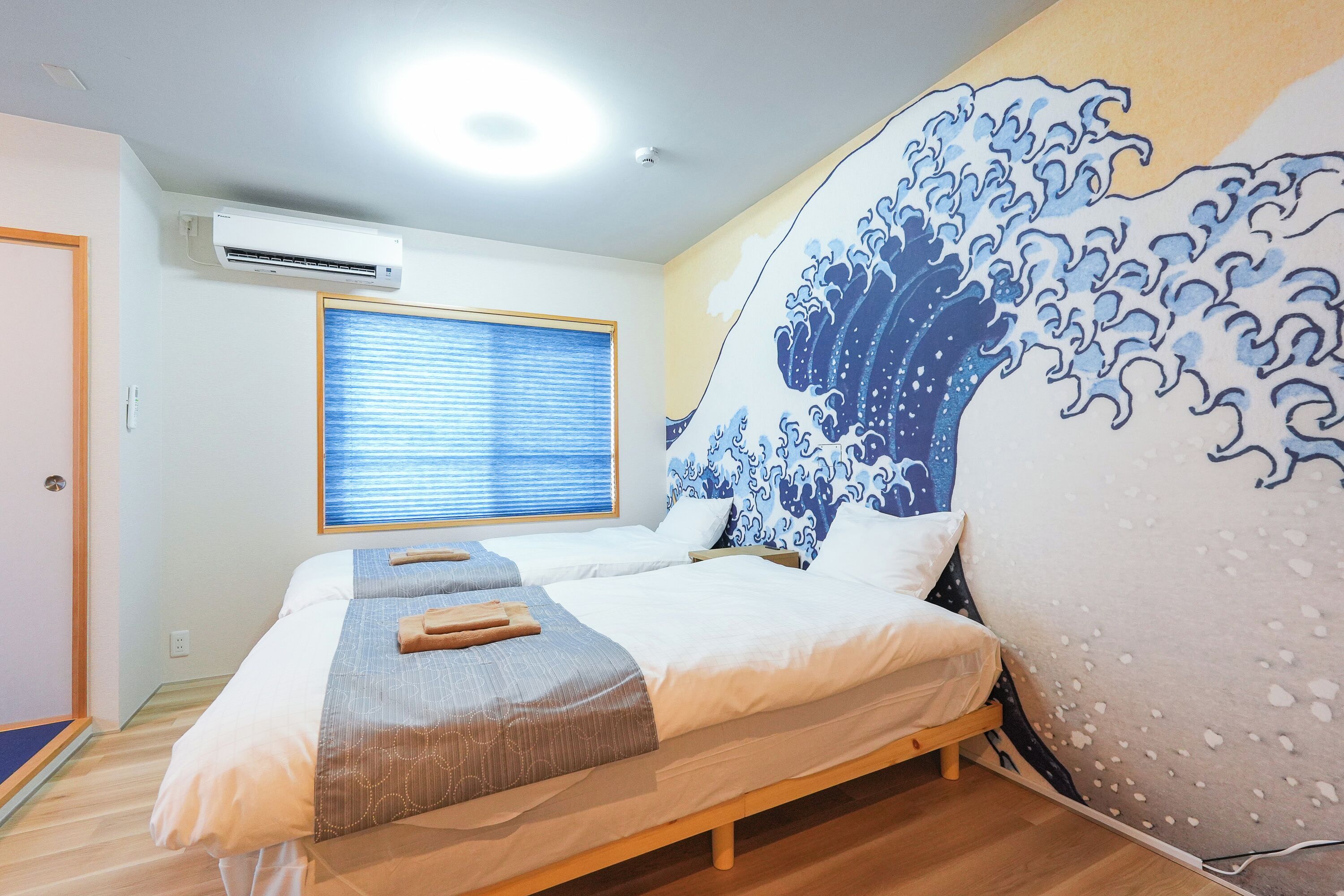 A hotel room with two beds, an air conditioner, a window with blinds, and a wall with a wave painting.