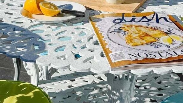 Uma mesa com toalha de renda branca, uma tigela, uma caneca e um prato com fatias de laranja.