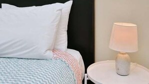 Uma cama com roupa de cama branca e estampada, cabeceira preta e mesa de cabeceira redonda branca com uma luminária.