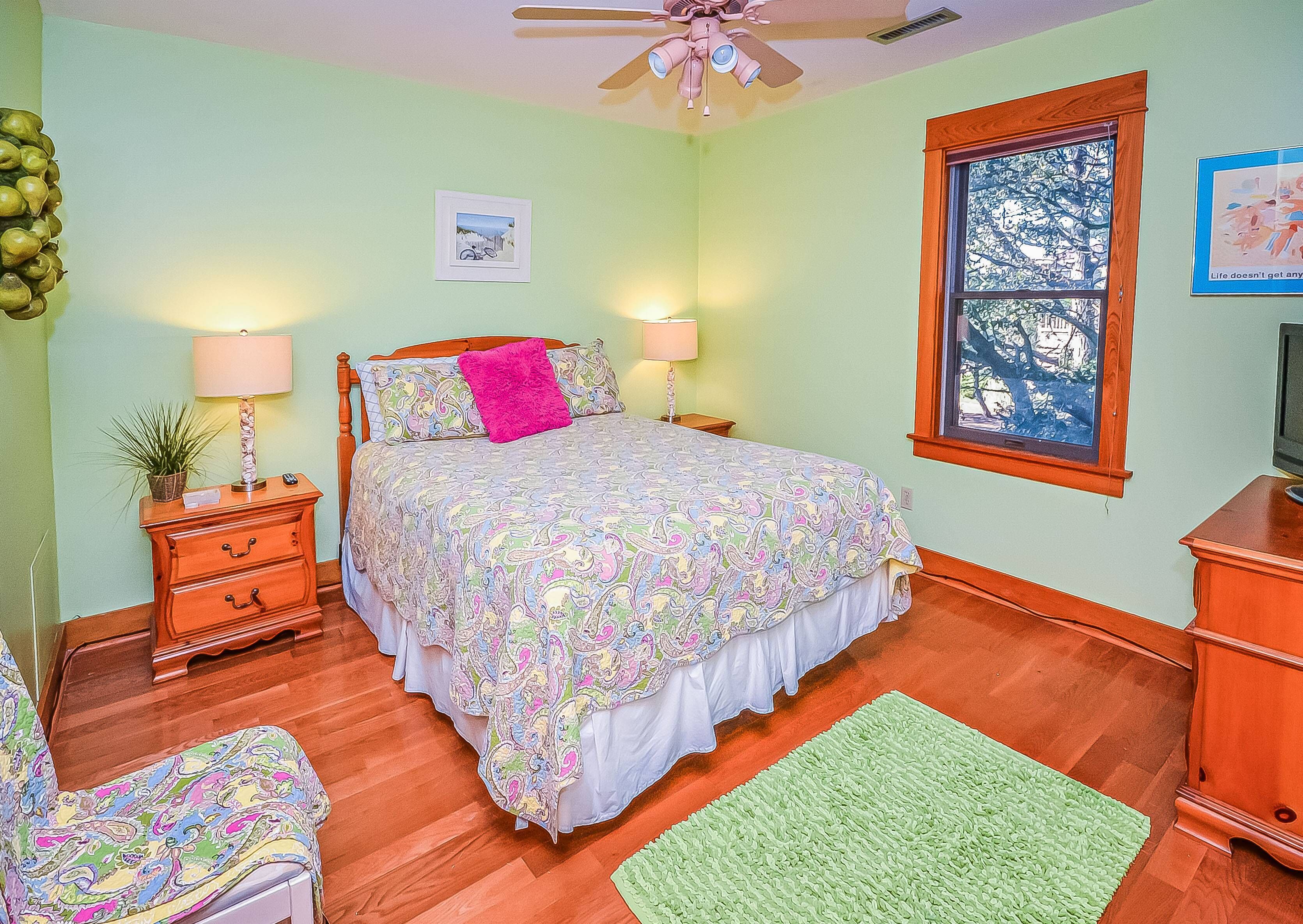 A bedroom with a bed, two bedside tables with lamps, a green rug, a wooden dresser, and a window with a view of trees.