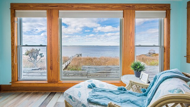 Ett sovrum med ett stort fönster med utsikt över en strand och havet, en takfläkt och en säng med ett blått täcke.