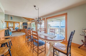 Un coin repas avec une table et des chaises en bois, un lustre et de grandes fenêtres donnant sur l’océan.