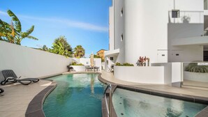 Una piscina amb gandules, un parasol i un edifici blanc amb façana corbada.
