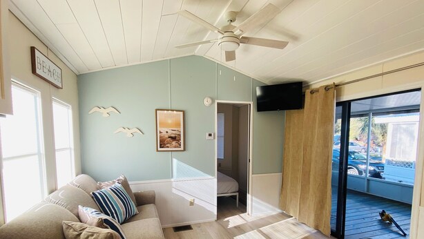 Una habitación con ventilador de techo, un sofá con cojines, una cama, un televisor montado en la pared y vistas al exterior a través de una puerta de cristal.