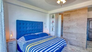 Een slaapkamer met een blauw gestoffeerd hoofdbord, een gestreept beddegoed en een nachtkastje met een lamp.