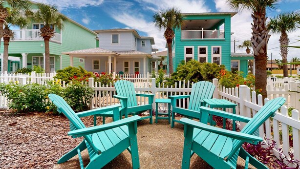Un bâtiment situé sur la côte, avec une clôture blanche en bois, des fauteuils Adirondack turquoise et un ciel d’un bleu limpide.