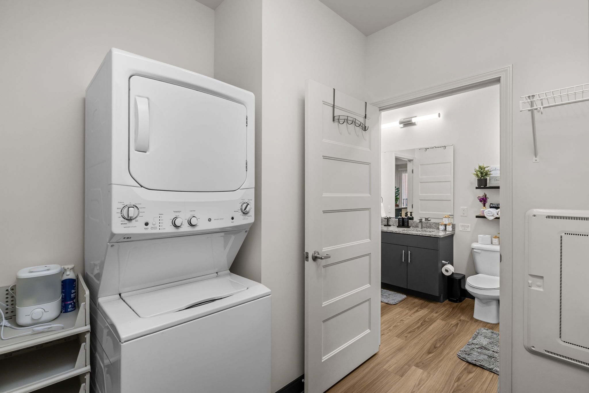 A laundry room with a front-loading washer and dryer, a door leading to a bathroom, and a small shelf with a bottle and a container.