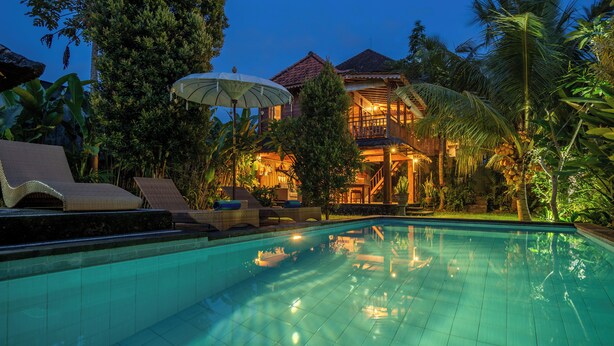 A swimming pool with lounge chairs and umbrellas, surrounded by lush greenery and a building with a red roof. - Aris Villa Ubud (Ubud)