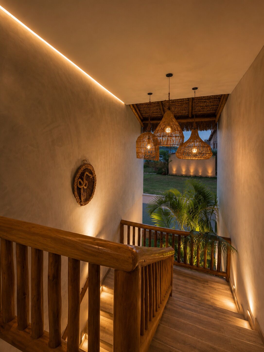 A wooden staircase with a railing, leading to a balcony with hanging wicker lamps and a view of greenery.