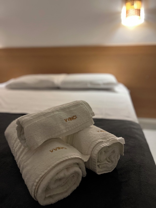 A neatly made bed with white and black bedding, a VINCIGALL towel, and a wall-mounted light fixture. - Villa Vinci Flat (São Miguel do Gostoso)
