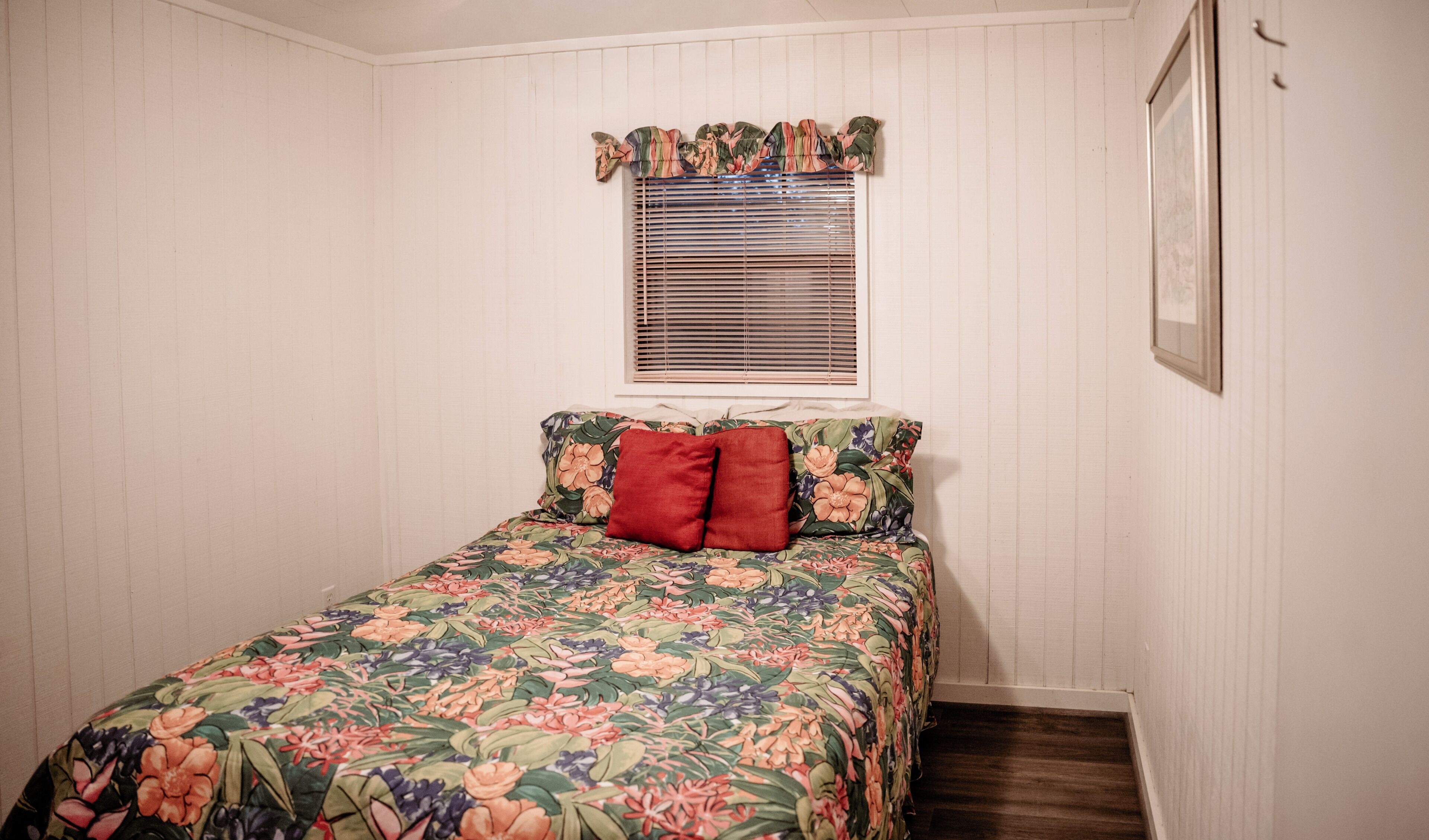 A bedroom with a floral bedspread, a window with blinds, and a framed picture on the wall.