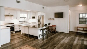 A modern kitchen with white cabinetry, a central island with stools, a ceiling fan, and a flat-screen TV mounted on the wall. - Riverfront, HotTub, BoatRamp, Deck, Sleeps 24 (West Columbia)