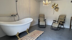 Ein modernes Badezimmer mit freistehender Badewanne, zwei schwarzen und beigefarbenen Stühlen, einer Holzbadeinmatte, einem Chandelier und Wandleuchtern in Form von Blättern.